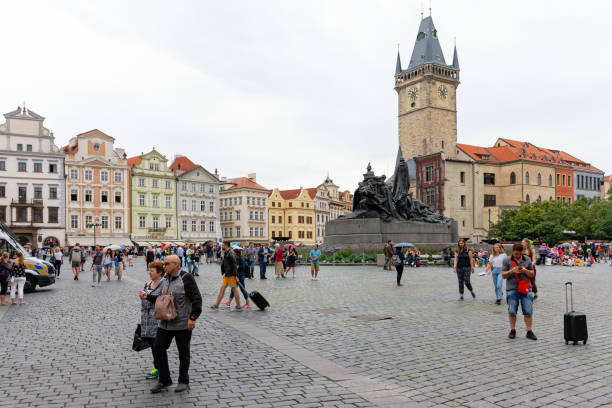 Prague, Bohemia, Czech Republic, august 10th 2018, large group of people (walking, standing, sitting, shopping) on the old townsquare in Prague on a summer day, the square is surrounded by typical historical buildings, each in their own specific (pastel) color - in the centre of the square is the Jan Hus memorial (Czech philospher) and on the right side the old town hall (Staroměstská radnice)\n\nPrague is considered to be one of the most beautiful cities in Europe and is a popular travel destination for tourists, the historic old town is one of the main tourist attractions and included in the UNESCO list of World Heritage Sites