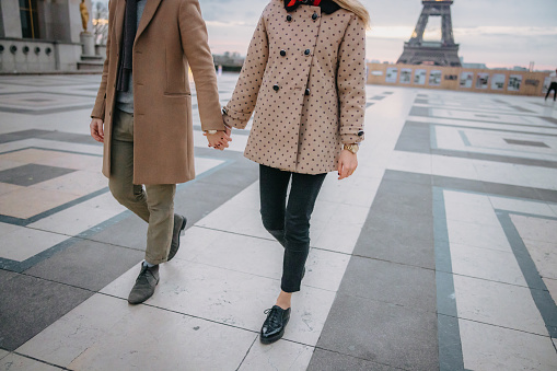 Young couple, man and woman wearing beige coat and jacket holding hands while walking on Parvis des Droits de l'Homme, focus on lower body parts, Eiffel Tower in background, focus on forefront, horizontal