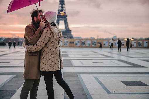 Young couple, woman with long blonde hair wearing a white hat and beige jacket with brown dots, man with short brown hair and beard, wearing a beige coat, kissing at Parvis des Droits de l'Homme, Paris in the evening in front of Eiffel Tower, man holding a pink umbrella, focus on forefront, horizontal
