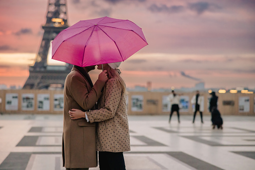 Couple, man with beard, wearing a beige coat, woman with long blonde hair wearing a white hat and beige jacket with brown dots, kissing and hugging while standing on Parvis des Droits de l'Homme in Paris during twilight, man holding a pink umbrella over both of them, people in the background and Eiffel Tower with beautiful sky, focus on forefront, horizontal