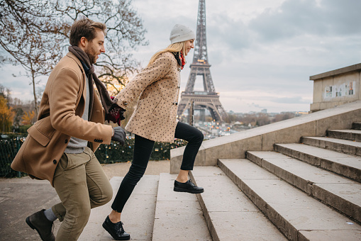 Young couple, woman with long blond hair wearing a white hat and beige jacket with brown dots, man with short brown hair and beard, wearing a beige coat, holding hands while running up stairs to the Parvis des Droits de l'Homme, Paris, Eiffel Tower in the background, focus on forefront, side view, horizontal