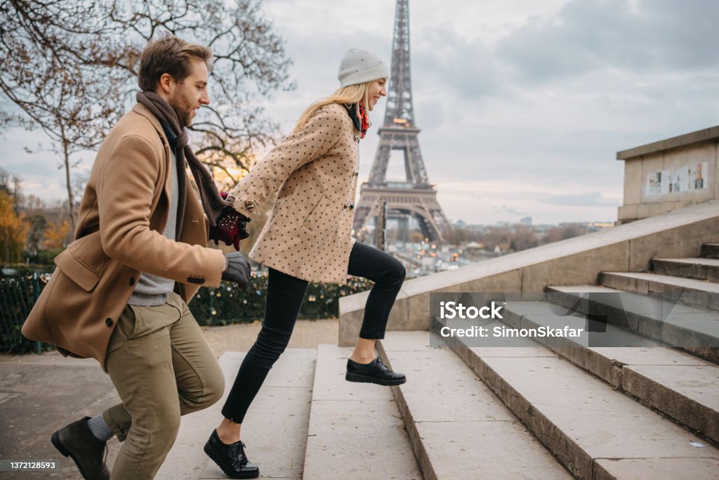 Young couple running up stairs to Parvis des Droits de l'Homme, Paris, in front of Eiffel Tower Young couple, woman with long blond hair wearing a white hat and beige jacket with brown dots, man with short brown hair and beard, wearing a beige coat, holding hands while running up stairs to the Parvis des Droits de l'Homme, Paris, Eiffel Tower in the background, focus on forefront, side view, horizontal Paris - France Stock Photo