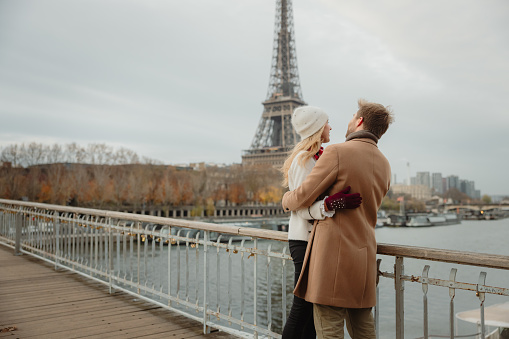 Romantic couple, woman with long blond hair wearing a white hat and white jacket, man with short brown hair and beard, wearing a beige coat, hugging each other closely while standing on a bridge at Seine River, Paris, man looking at his girlfriend, girlfriend looking at the river, upper body parts, Eiffel Tower in background, horizontal