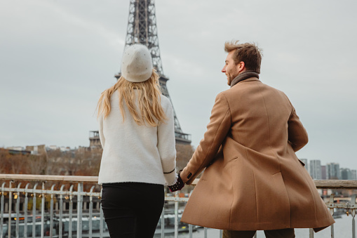Couple, woman with long blond hair wearing a white hat and white jacket, man with short brown hair and beard, wearing a beige coat, are excited to see Eiffel Tower, Paris, the first time at their France holiday, man looking at his girlfriend both holding hands, rear view, horizontal