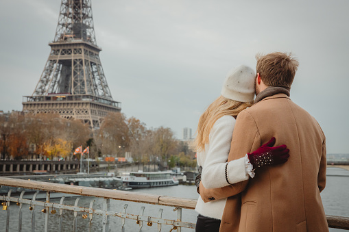 Couple, woman with long blond hair wearing a white hat and white jacket, man with short brown hair and beard, wearing a beige coat, standing on a bridge at Seine River, hugging each other closely while looking at Eiffel Tower, Paris, rear view, upper body parts, horizontal