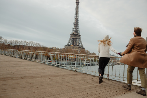 Woman with blond hair, wearing a white hat and white jacket holding hands with her boyfriend while running on a bridge at seine river to see Eiffel Tower Paris, rear view, cloudy day, horizontal