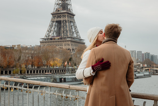 Romantic couple, woman with blond hair, wearing a white hat and white jacket kissing her boyfriend on the cheek while both are hugging in front of Eiffel Tower, Paris, standing on a bridge at Seine River. Upper body parts, horizontal