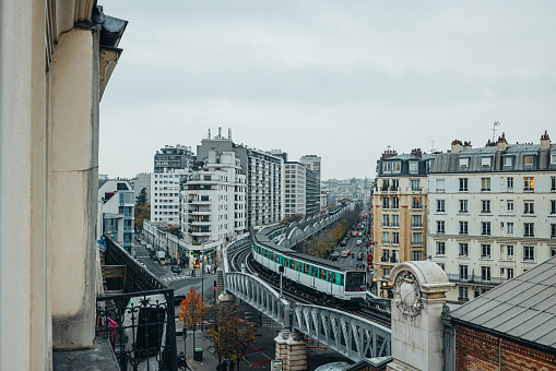 Metro train with white and green color is driving through the center of Paris during a cloudy day, plenty of building facades around the train track, metro above ground on a bridge, horizontal