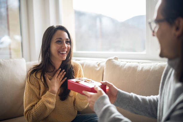 Cheerful young woman receiving a gift from her boyfriend. Boyfriend surprise his beautiful girlfriend with present while she is sitting on the sofa in the living room at home. gift for girlfriend stock pictures, royalty-free photos & images