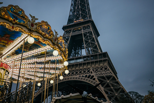 Eiffel Tower in the evening without lighting, carousel with plenty of light bulbs and lighting in forefront, view from low angle, cloudy sky in the background, horizontal