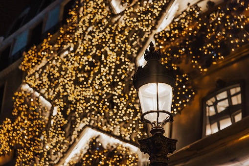 Old traditional French street light in Paris with lighting during nighttime, hundreds of Christmas light bulbs on a house facade in the background, low angle view, horizontal