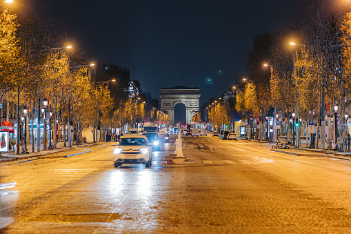 Avenue des Champs-Elysees with moderate car traffic on the street during night time, trees and orange street lights next to the street, Arc de Triomphe in the with beautiful blue night sky in the background, horizontal