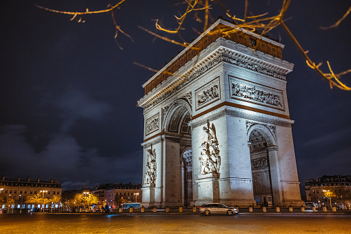 Arc de Triomphe with lighting during nighttime with beautiful cloudy night sky, no car traffic around the monument just one car driving in front of the building, low angle view, branches of a tree in forefront, horizontal