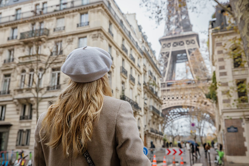 Blond Woman, wearing a white hat and beige coat, crossing the street on a crosswalk, walking in the direction of Eiffel Tower, Paris during daylight, looking at the tower, facades of old traditional buildings in the forefront of the Tower, rear view, horizontal