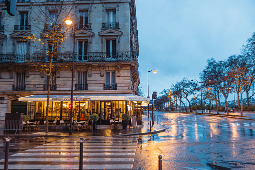 Beautiful traditional French cafe in the corner of a side street in Paris, cafe is closed with chairs on the table, rainy day with wet street in front of the cafe, trees with beautiful early morning sky in the background, horizontal