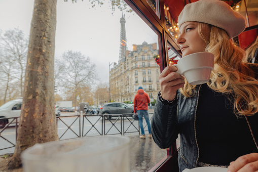 Woman with blond hair, wearing a white hat and black leather jacket, sitting in a traditional French cafe in Paris, looking outside the window, rainy outside, wet sidewalk and street, holding a cup of Coffee, Eiffel Tower in the background, horizontal