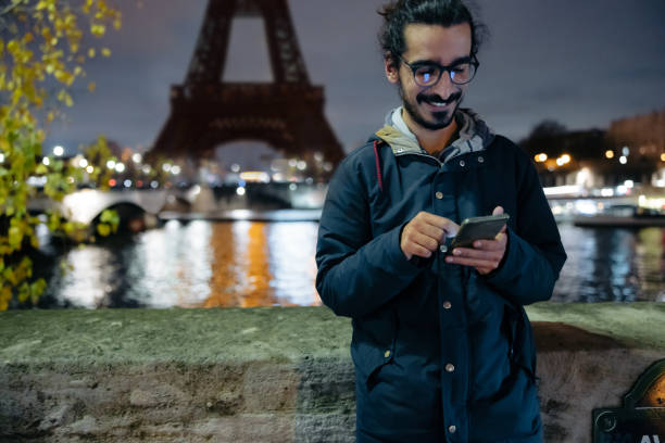 Man with black hair tied in a braid and beard, wearing blue jacket and glasses, smiling while looking down at his smartphone in the evening, leaning against a concrete wall at seine river, Eiffel Tower, Paris with lighting on the other side of the river in background, upper body parts, focus on forefront, horizontal