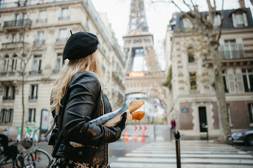 Blond Woman, wearing a leather jacket and black hat, standing at a crosswalk, carrying two French baguettes under her right arm, two old traditional Buildings with Eiffel Tower, Paris in the background, focus on forefront, rear view, early in the morning, low angle, horizontal