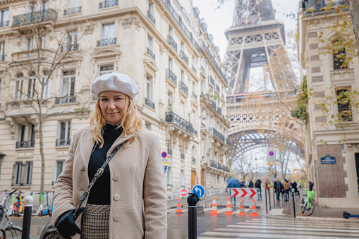 Blond adult Woman, wearing a beige coat and a white hat, looking at camera while standing in front of Eiffel Tower, Paris, Crosswalk and traditional old Building facades in the forefront of the Tower, low angle view, upper body parts, horizontal