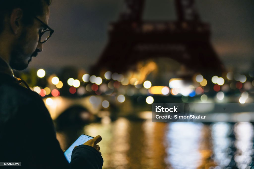 Close-up of man looking down at his Smartphone in front of Eiffel Tower, Paris, in the evening Close-up of man with black hair and beard, wearing blue jacket and glasses, looking down at his smartphone while standing on the other side of seine river in front of Eiffel Tower, Paris with lighting, in the evening, horizontal Paris - France Stock Photo