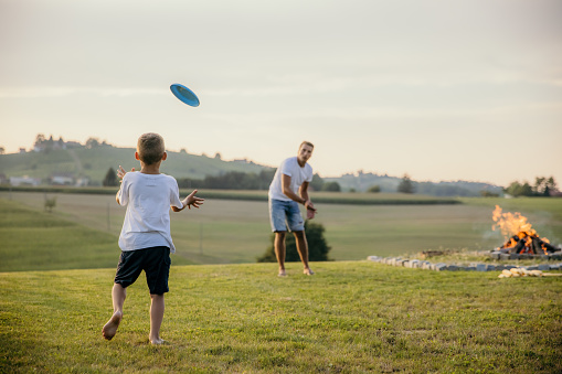 Close-up father with short brown hair, wearing a white t-shirt and jeans shorts, plays Frisbee with his little son with short blond hair, wearing a white t-shirt and black shorts on a meadow during a campfire in the summer, both of them are barefoot, twilight with sunset sky in the background, horizontal