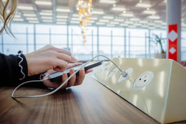 Close-up of a woman with blond hair, wearing a black pullover, using her smartphone while charging it via a white USB cable on a public charging station installed on a wooden table inside an airport terminal, window of the terminal in the background, focus on forefront, horizontal