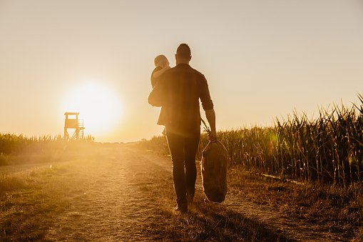Farmer, adult man with short brown hair, wearing a red checkered shirt and jeans, carrying his little blond haired son in his left arm and a bag of corn plant leaves in the right hand, walks on a country lane during sunset, raised hide in the background, clear sky, backlight, rear view, horizontal