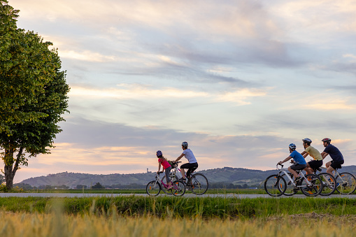 Family, parents with two Sons and one daughter biking on a country road in the evening, all of them wearing shorts, t-shirts and helmets, mother and daughter in the forefront looking at the hills on the horizon, father riding with his sons behind them, low angle view, side view, horizontal