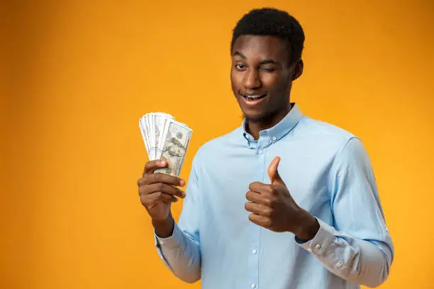 Photo of Excited black guy holding stack of Dollars in yellow studio
