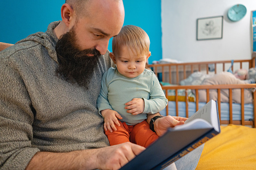 Caucasian father reading a book, while his curious toddler son listen to him