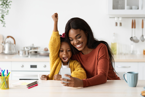 Happy Excited Black Mother And Little Daughter Celebrating Success With Smartphone While Sitting At Table In Kitchen, Cheerful African American Mom And Female Child Playing Video Games At Home