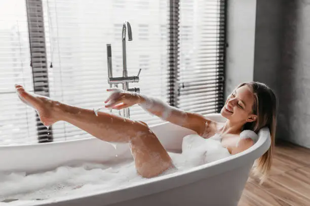 Personal care at home. Happy lady shaving legs with razor, lying in foamy bubble bath, doing skin care procedure. Home wellness and beauty, hair removal, depilation concept