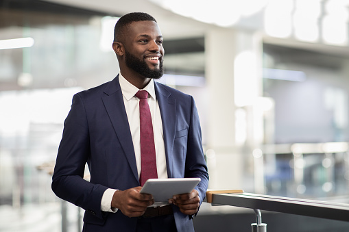 Cheerful young black bearded man manager walking by office building corridor, holding modern digital tablet, using mobile app for business, looking at copy space and smiling, panorama