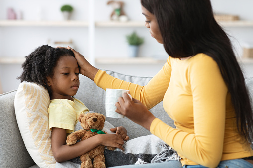 Caring Black Mother Giving Hot Tea To Her Ill Little Daughter And Touching Her Forehead, Loving African American Mom Taking Care About Sick Female Child Suffering Seasonal Flu Or Cold, Closeup