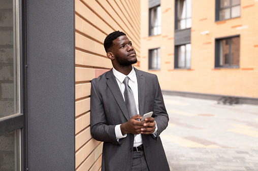 Strategic thinking. Thoughtful african american businessman holding smartphone and looking away, leaning on wall, standing near office building, free space
