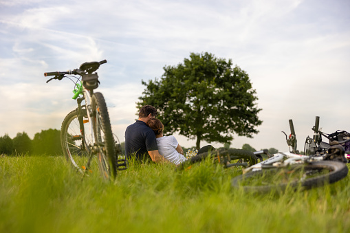 Parents taking a rest from their bike trip, sitting closely and hugging on a meadow during summer, woman leaning her body against her husband, bikes of the entire family lying in the forefront, a bike standing behind them, beautiful tree and sky in the background, low angle, rear view, horizontal