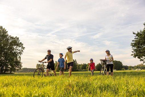 Parents pushing their bikes on a meadow while their children, two sons and a daughter, walking next to them, oldest son is pointing with his hand on something while looking away, his brother and sister walking between the parents, all of them wearing short summer clothes, meadow in the forefront, trees, haystack and sky in the background, front view, horizontal