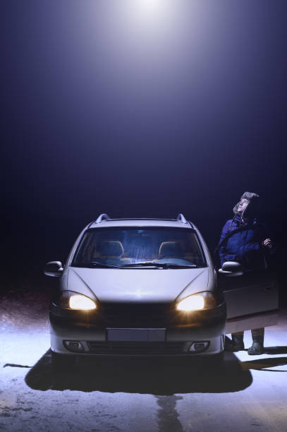 The man stands next to the car and looks up at the beam of light coming from the UFO. Dark winter night. Vertical stock photo