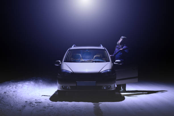 The man stands next to the car and looks up at the beam of light coming from the UFO. Dark winter night. Colored in Very Peri trend color stock photo