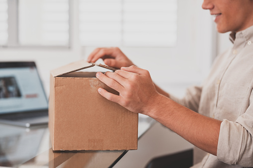 Smiling man wearing glasses unpacking awaited parcel, looking inside, sitting at work desk, satisfied happy customer opening cardboard box with online store order, good shipping delivery service