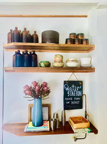 Horizontal still life interior of wood shelves in cafe with handmade ceramic bowls bottles and flowers in vase next to a drinking water filter tap in rural country cafe Bangalow near Byron Bay NSW Australia