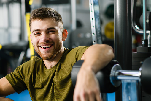 Portrait of smiling handsome young fit man with beard in gym interior with modern equipment machines, healthy active lifestyle
