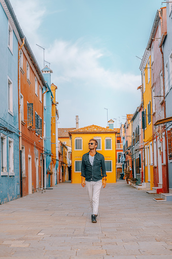 Portrait of a happy young handsome traveller man with sun glasses and sitting, smiling, having fun outdoor and walking against colourful houses on the canal in famous travel destination Burano island and boats lining side canals with beautiful reflection in Burano, Venezi, Veneto, Italia