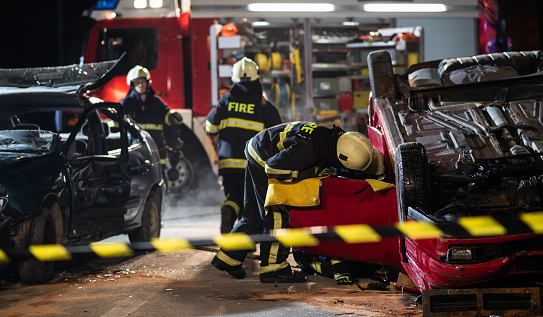 Night shot of three firefighters, one is carrying fire extinguisher, other two are opening door on upside down car, fire engine in background. Saving lifes and saftey concept.