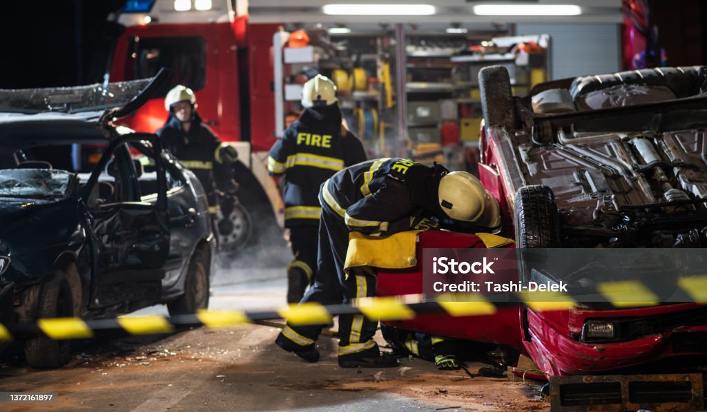 Firefighters At A Car Accident Scene Night shot of three firefighters, one is carrying fire extinguisher, other two are opening door on upside down car, fire engine in background. Saving lifes and saftey concept. Car Accident Stock Photo