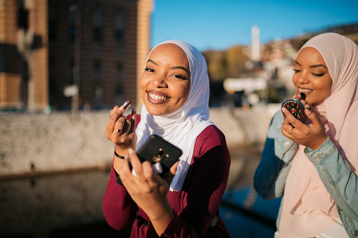 Portrait of two happy Muslim women wearing a hijab, sitting next to a river in the city, putting on a makeup and smiling