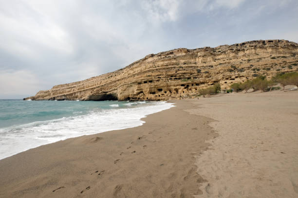 Matala, beautiful beach on Crete island, waves and rocks. Panorama Matala beach, Greece Panorama of Matala beach with the caves on the rocks that were used as a roman cemetery and at the decade of 70's were living hippies from all over the world, Crete, Greece crete cartoon stock pictures, royalty-free photos & images