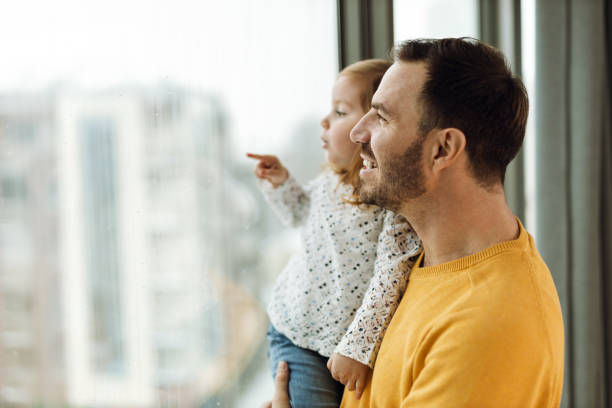 Happy father and his small girl looking through window from their home
