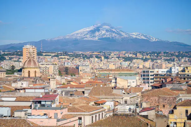 Skyline of Catania city center from Saint Agatha church terrace, with historic buildings and church cupolas, and Mount Etna volcano erupting in Sicily, Italy