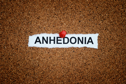 A crumpled piece of paper with the word Anhedonia on it pinned to a corkboard. Close up.
